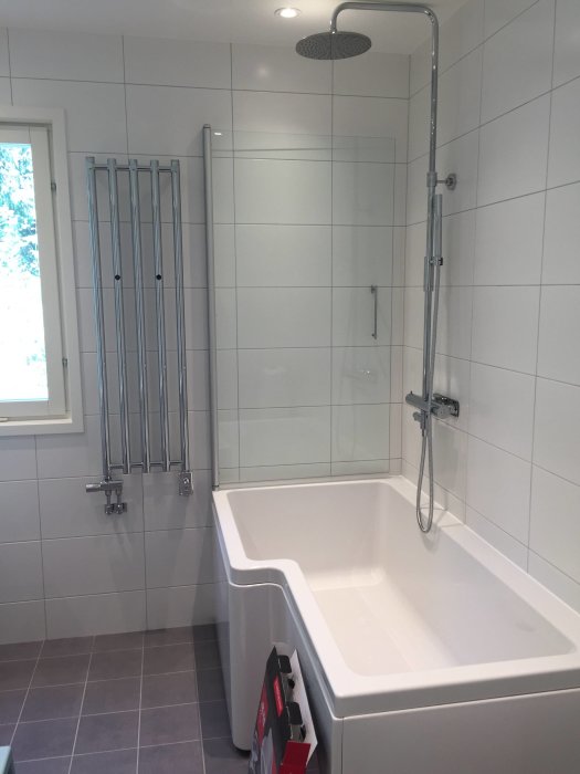 Svedbergs badkar med enkel duschvägg, takdusch och handduksradiator i vitt kaklat badrum.