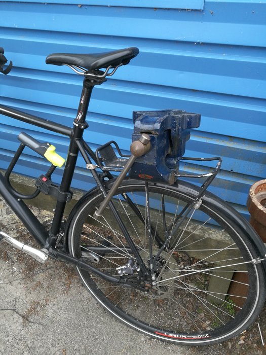 Cykel parkerad vid blå vägg med fastsatt äldre bänkskruvstäd på pakethållaren.