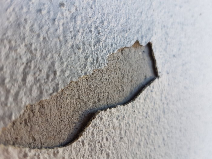 Närbild på fasad där ett elastiskt ytterlager har börjat släppa från underliggande puts på hålsten.
