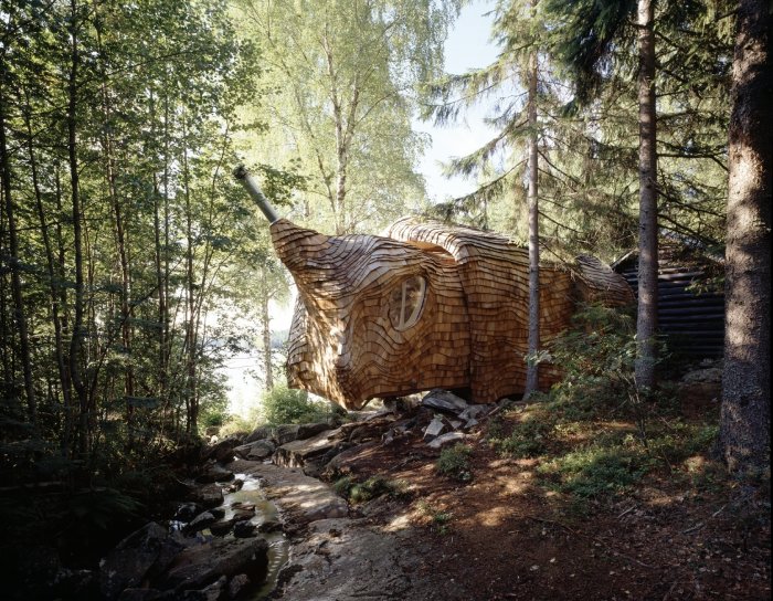 En byggnad formad som en gigantisk träsnigare i en skogsmiljö med träd runtom.