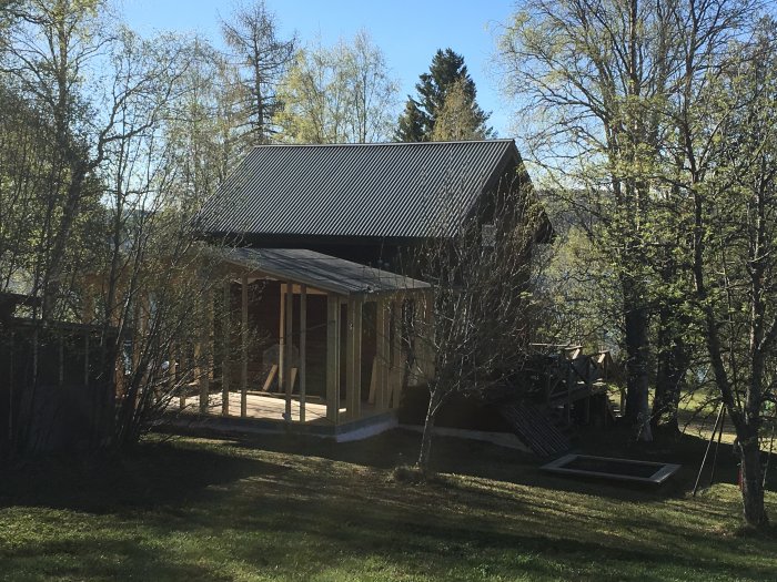 Nybyggt trähus med nästan färdiglagt takpapp under solljus, omgivet av gröna träd.