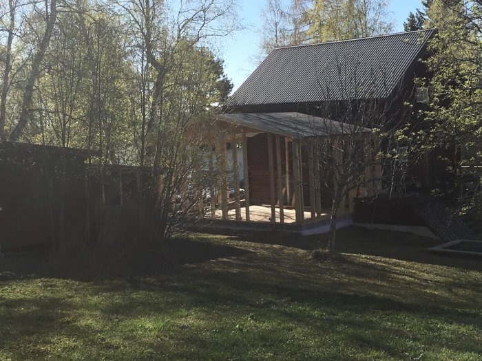 Byggprojekt med nylagt takpapp på en konstruktion nära en befintlig byggnad med träd och grön gräsmatta i förgrunden.