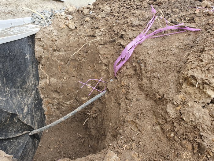 En elkabel synlig i en grävd gräva nära en byggnadsvägg, markerad med lila band, belägen på ca 300 mm djup.