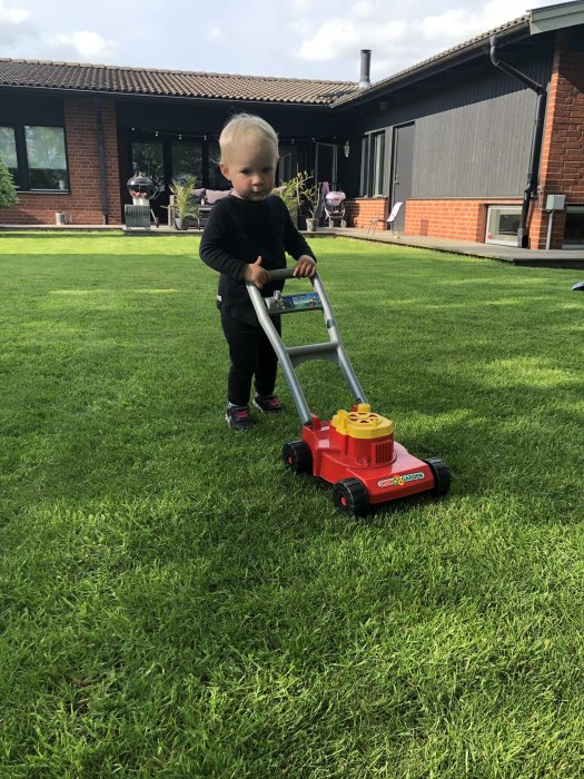 Barn leker med en leksaksgräsklippare på en välskött gräsmatta framför ett hus.