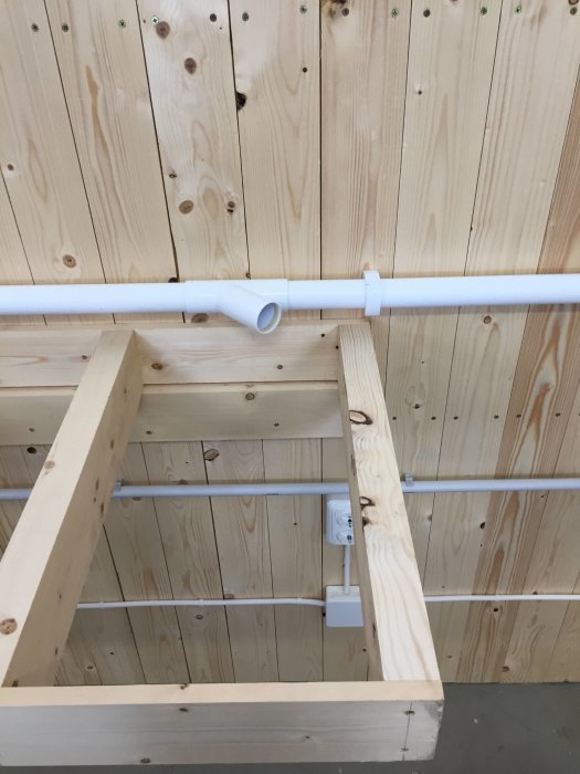 En träklädd vägg i ett garage med en elkontakt och en vit PVC-rörinstallation för dammutsug ovanför en träbänk under konstruktion.