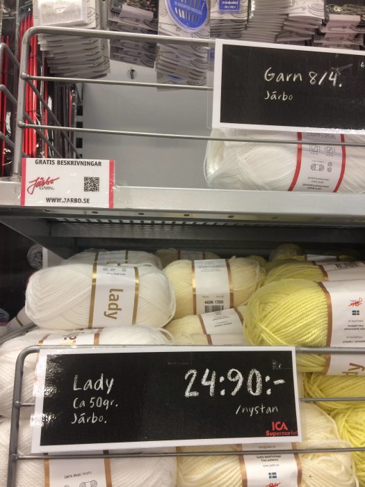 Hyllor med nystan av vitt och gult garn märkta 'Lady' och 'Apli' med priset 24:90 i en butik.