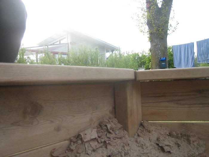 Inblick i en hembyggd, sexkantig sandlåda med träplankor och sand, med utsikt mot trädgård och tvätt på tork.