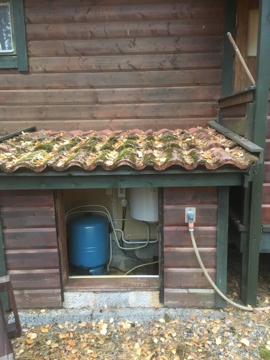 Ett litet pumphus med öppen dörr, visande installationer och ett blått tryckkärl inuti.