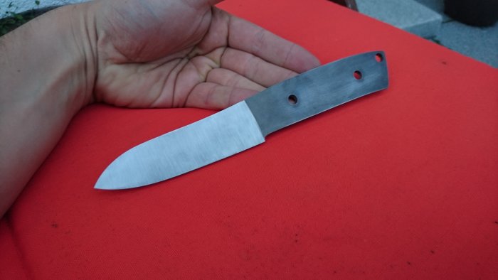 Hand håller ett slipat knivblad med Loveless-design och fårfot, i stål RWL 34, ovanpå rött tyg.