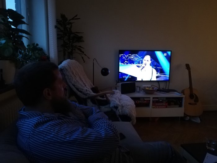 Man sitter i soffa och ser fokuserat på TV där en person framträder på scenen, med en gitarr i förgrunden.
