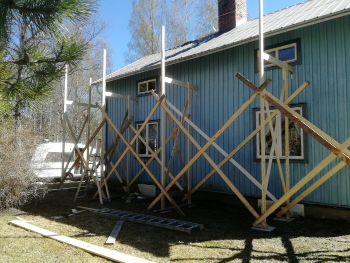 Träställning konstruerad intill ett blått hus med syfte för tak- och fasadrenovering under en solig dag.