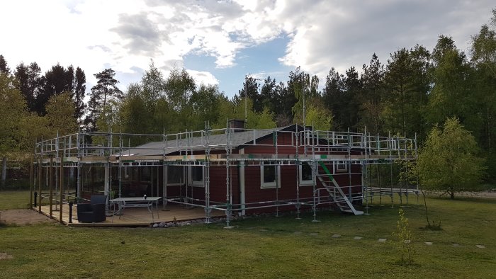 Rött hus från 1963 omgivet av byggställningar under renovering av tak, med trädgård och träd i bakgrunden.