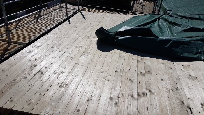 Ny råspont på tak med delvis täckning av byggpresenning, i solljus.