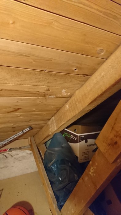Oisolerat vindsutrymme med synliga träbjälkar och lutande tak, delvis fylld med föremål som kartonger och en basketball.