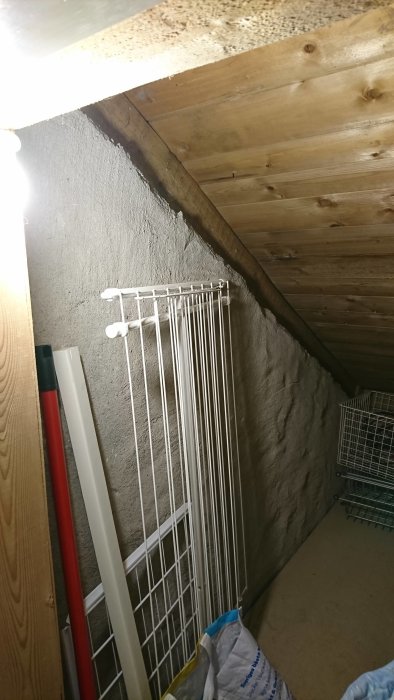 Oisolerad kattvind med synliga träbjälkar och betongvägg, oordning och förvaring.