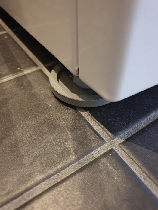 Tvättmaskinsfot placerad på en dämpare som ligger på ett mörkt kakelgolv.