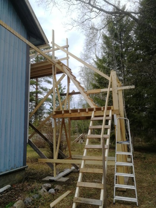 Nybyggt trapphus i trä vid sidan av ett blått hus med en stege lutad mot konstruktionen.