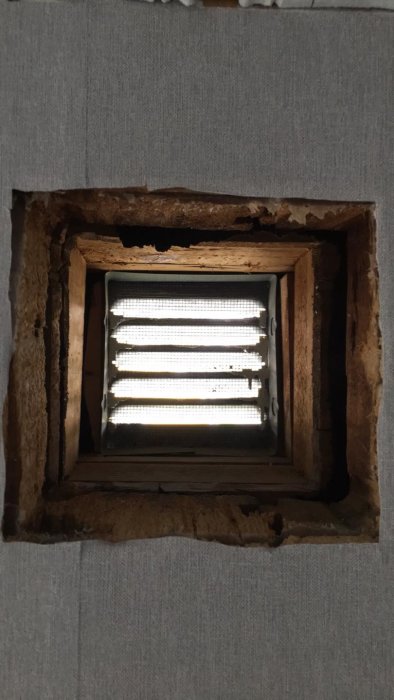 Vy inifrån av ett öppet ventilationsschakt i en vägg som avslutas med ett galler, omgivet av isolering och träväggsmaterial.