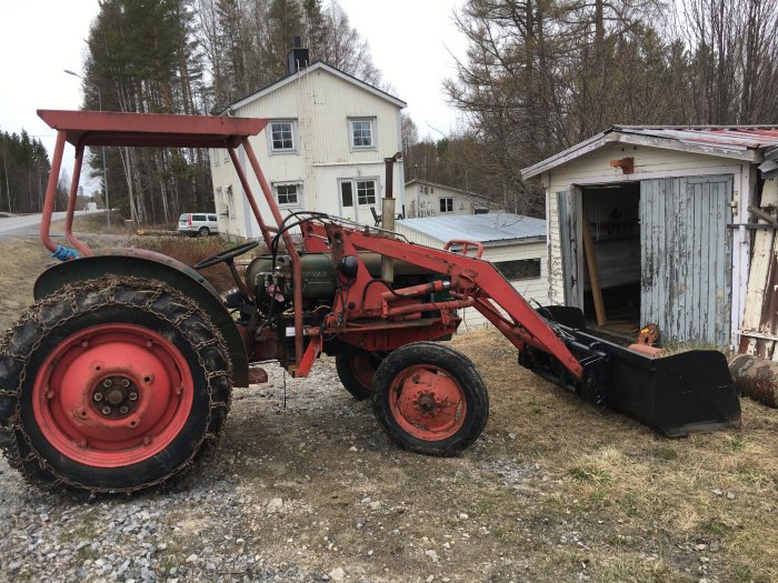 Gammal röd traktor med lastare framför vit byggnad, nära öppen skjul och träd i bakgrunden.