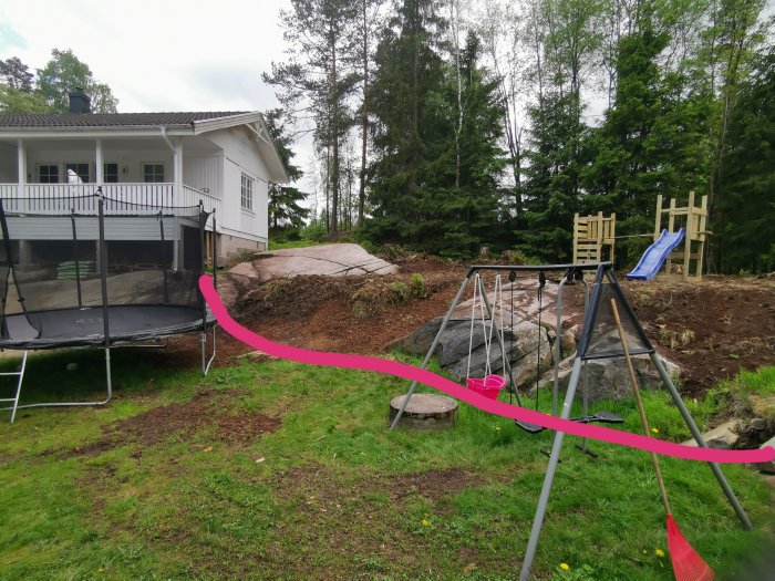 Trädgård med gräsmatta, en studsmatta, lekställning och exponerat berg, avsedd för framtida stödmur.