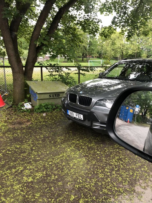 Grå BMW parkerad på gräs nära en sandlåda med löv på marken, sett från ett bilfönster.