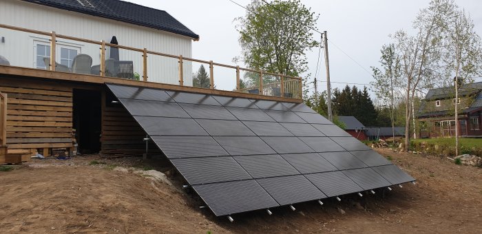 Solpaneler installerade på markstöd framför ett hus med trädäck.