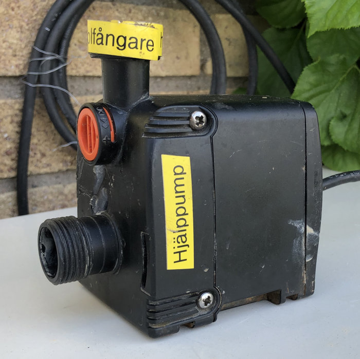 Svart liten booster-pump märkt 'Hjälppump' för solfångare med gula etiketter och kopplingsgänga framför tegelvägg.
