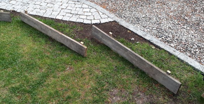 Träramar som markerar växtbäddar längs en kantstenad gångväg och grusad yta, med gräsmatta i förgrunden.