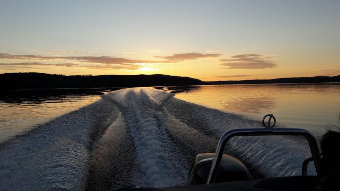 Solnedgång sett från en motorbåt på en sjö med skogklädda kullar i bakgrunden.