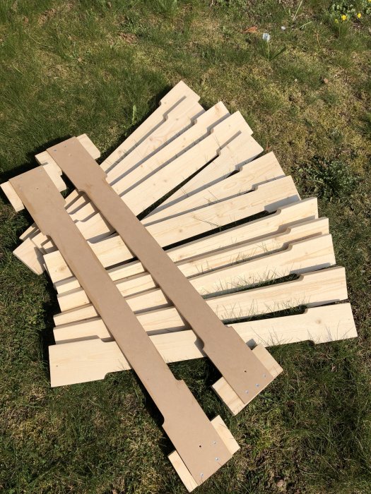 Hemgjorda träspjälor för räcke bredvid MDF-mallar på gräsunderlag under soligt väder.