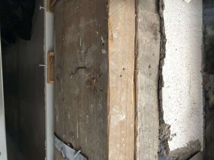 Två träbalkar stöttar en H-balk, synliga vid en öppning i väggen, bredvid isoleringsmaterial.
