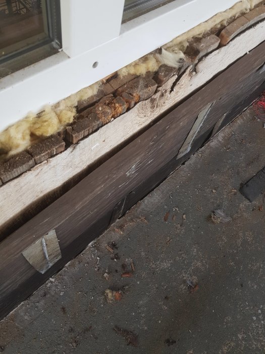 Närbild av ett gammalt fönsterkarmens nedre del visar slitage och isoleringsmaterial i ett hus från 1850.