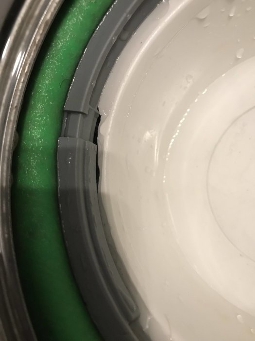 Närbild på en vit golvbrunn med synlig skada på kanten och vattendroppar, intill grön rörelselast.
