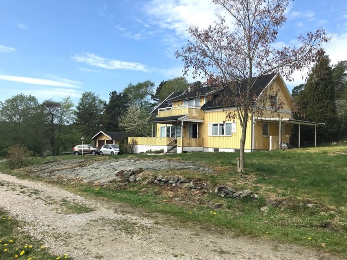 Gult hus med papptak och veranda i soligt, bergigt landskap, nära vatten, omgivet av träd.