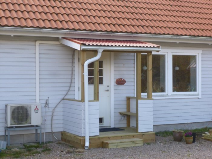 Nybyggd trätrapp framför husentré med tak, vit ytterdörr och fönster, bredvid värmepump.