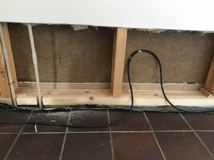 Vägg med borttagen panel som exponerar Asfaboard och reglar, bredvid en kabel på ett tegelgolv.