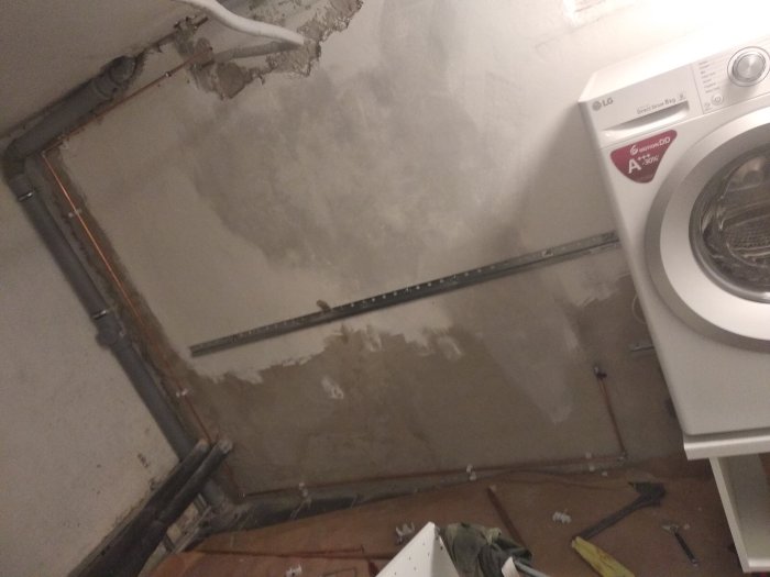 Oavslutad rörinstallation bredvid två tvättmaskiner i ett renoveringsprojekt, verktyg och rördelar synliga på golvet.