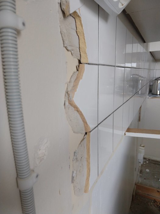 Delvis riven vita kakelväggen som visar tjockt lager av fix på gipsväggen bakom, med synliga rörledningar.