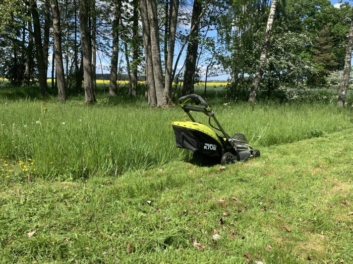 En elektrisk gräsklippare på en delvis klippt äng med träd och en rapsfält i bakgrunden.