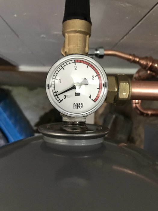 Manometer med visare på cirka 1,5 bar monterad på en grå vattentank med kopparledningar.