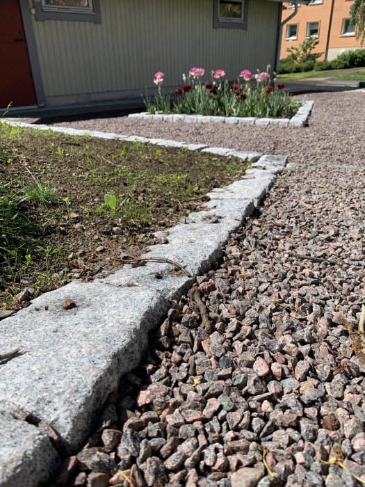 Stenläggning bredvid gräsmatta med blomrabatt i bakgrunden intill ett hus.