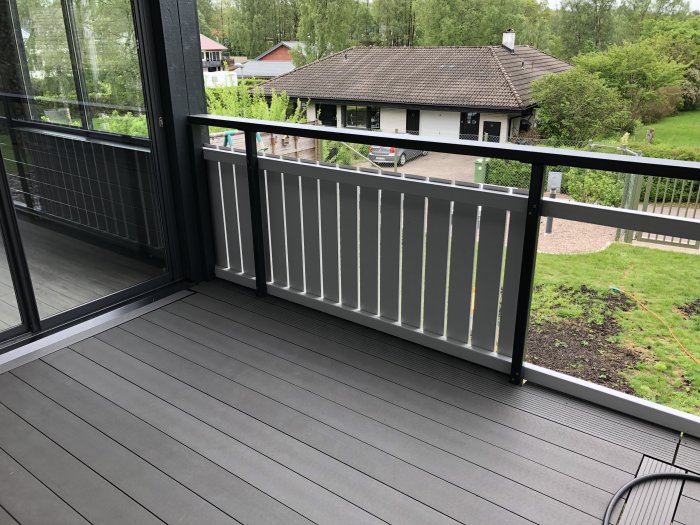 Delvis installerat vitt staket på en mörkgrå altan med utsikt över en trädgård och ett hus i bakgrunden.