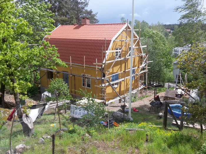 Hus under renovering med ställningar, gult trähus med rött tak i lummig trädgård.