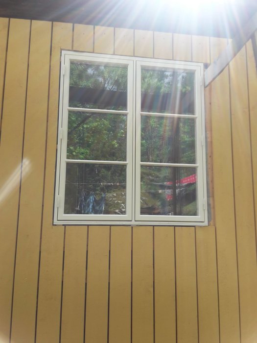 Vitfönstrad tvådelad fönster på en gul husvägg med solstrålar som speglar genom glaset.