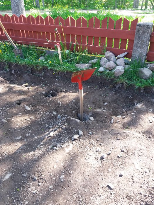 En spade i en uppgrävd jordyta med en röd staketbakgrund och synliga stenblock.