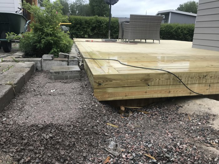 Nybyggd träterrass vid hus, med trädgårdsslang ansluten, bredvid grusgång och buskage, utomhus i dagsljus.