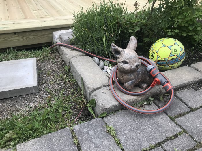 Slitna betongstenar, en kaninformad vattenspridare kopplad till en orange slang, en färgglad boll och träaltan i bakgrunden.