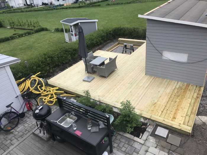 Vy över en halvfärdig uteplats med omonterad grill och gul vattenslang vid en trädgård.