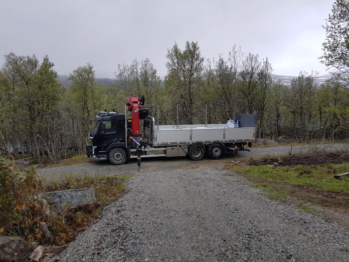 Kranbil med virkeslast parkerad på en grusväg med skarp sväng, omgiven av skog i snöfall.