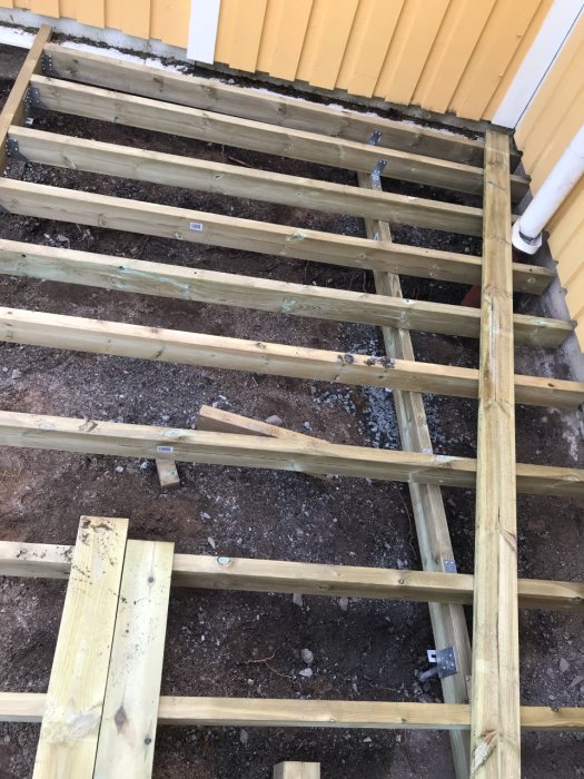 Byggnation av en träaltan med 120x45 tvärbalkar och metallbeslag nära en gul husvägg med överhäng.